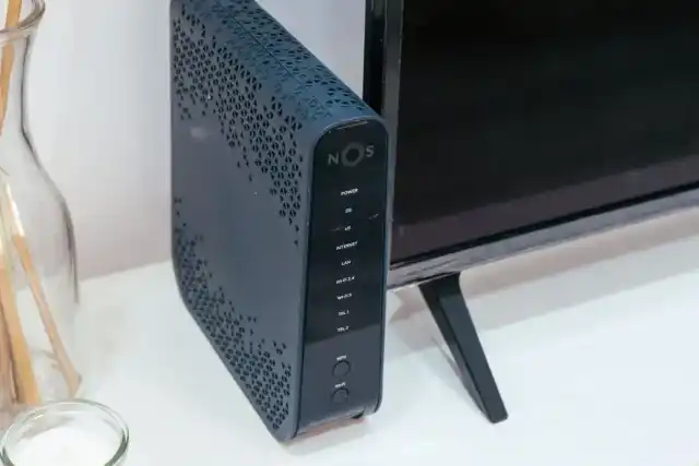 Roteador Wi-Fi sobre móvel na sala, ao lado de uma televisão e de objetos decorativos