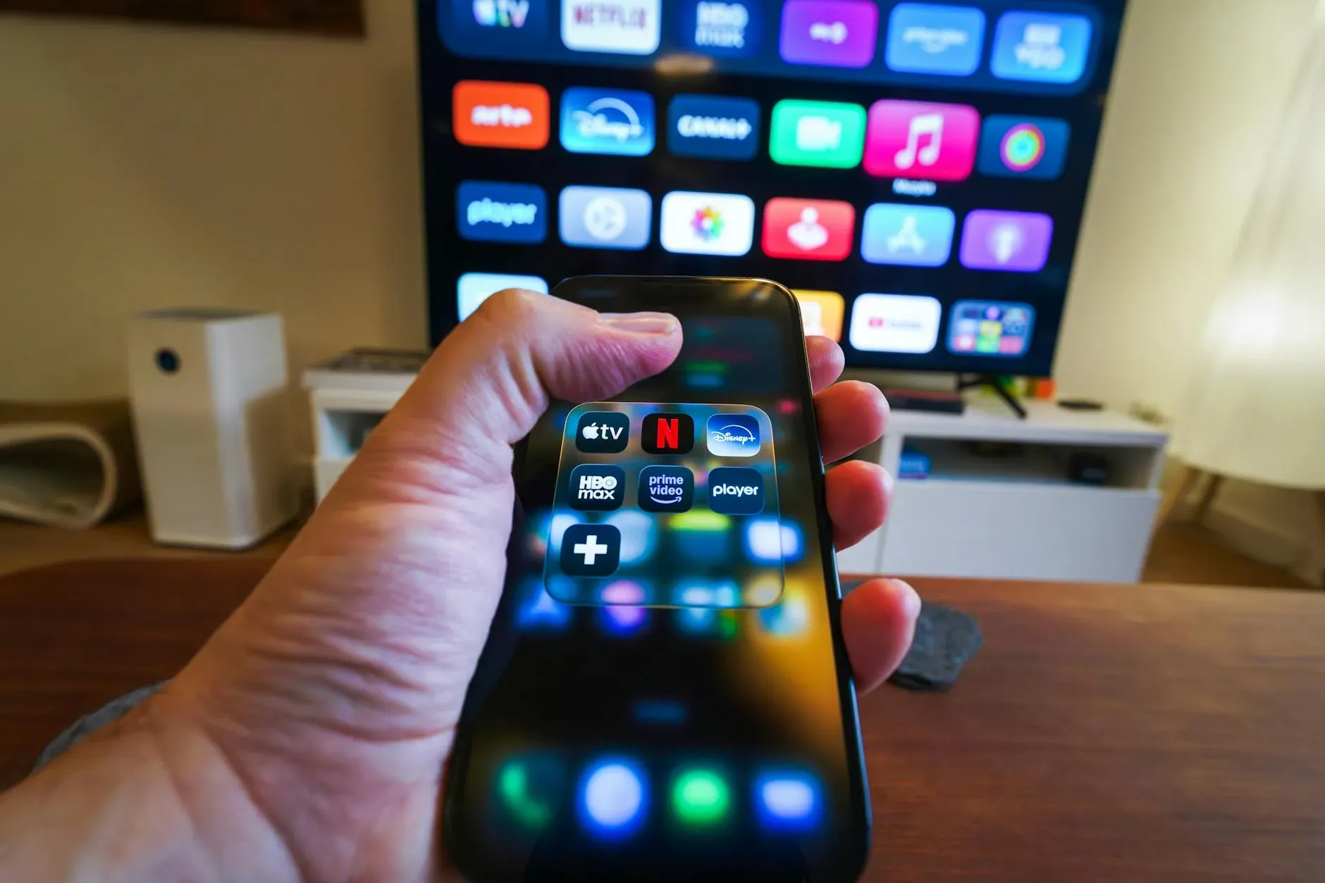 Imagem em close-up de uma mão segurando um celular com aplicativos de streaming exibidos em frente a uma TV com vários ícones de aplicativos.