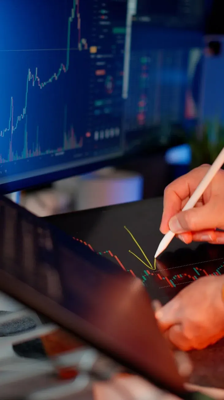 Pessoa usando uma caneta stylus em um tablet para análise do mercado de ações, com gráficos em um monitor.